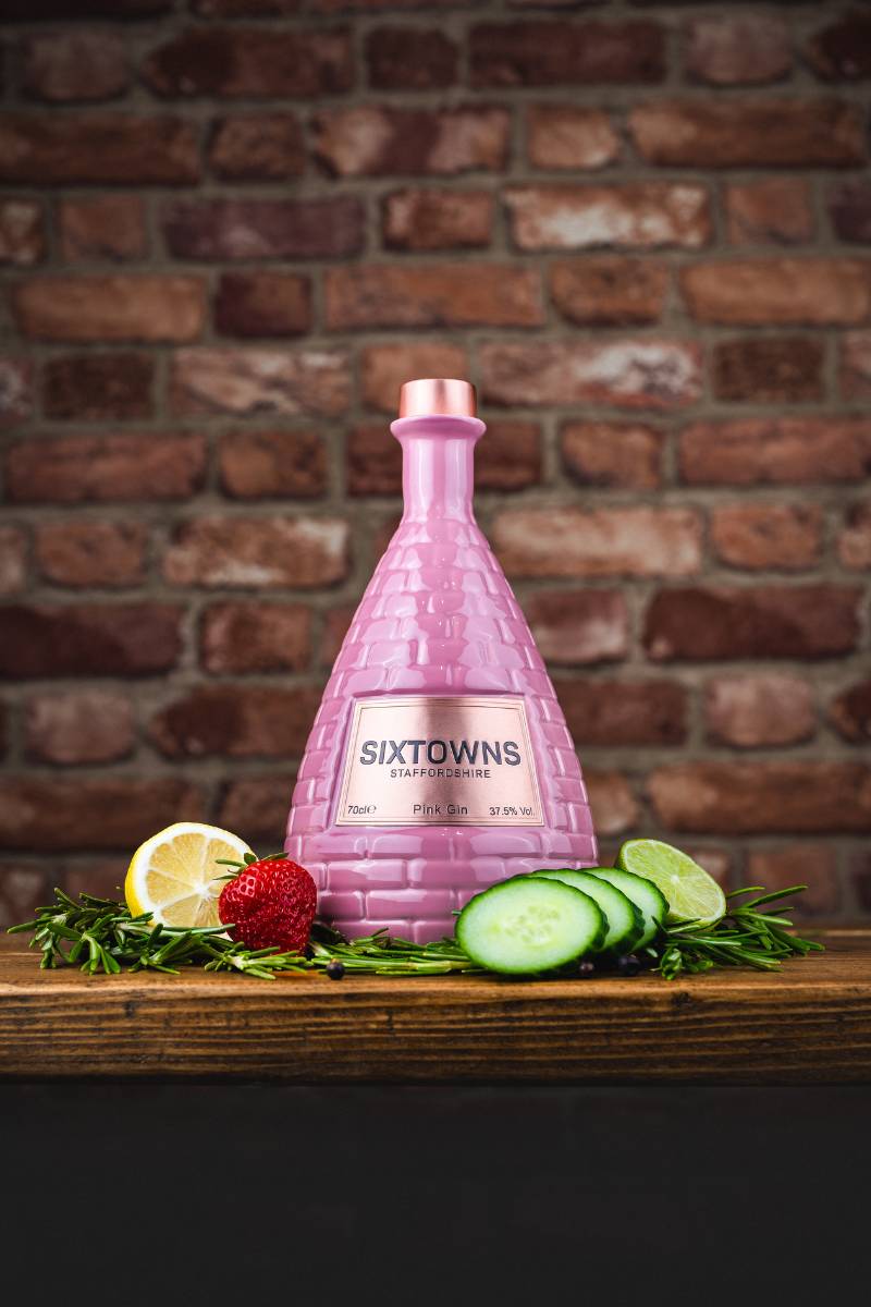 SIXTOWNS Pink Gin ceramic bottle on a wooden surface with strawberry, cucumber, lime, and lemon garnish, set against a rustic brick wall backdrop.