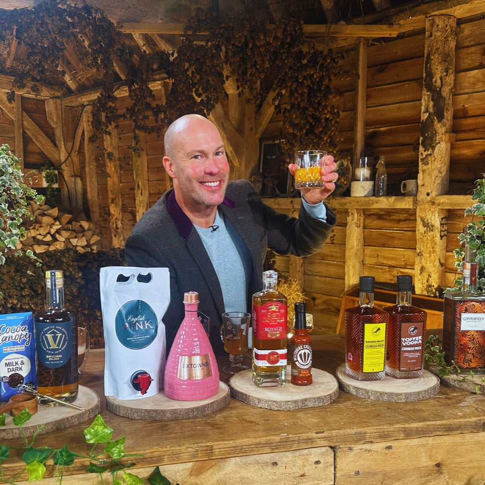 TV presenter smiling and holding a glass of spirit during an ITV showcase featuring a range of British craft spirits, including SIXTOWNS Pink Gin in a ceramic bottle, set against a rustic wooden bar with greenery and artisanal products on display