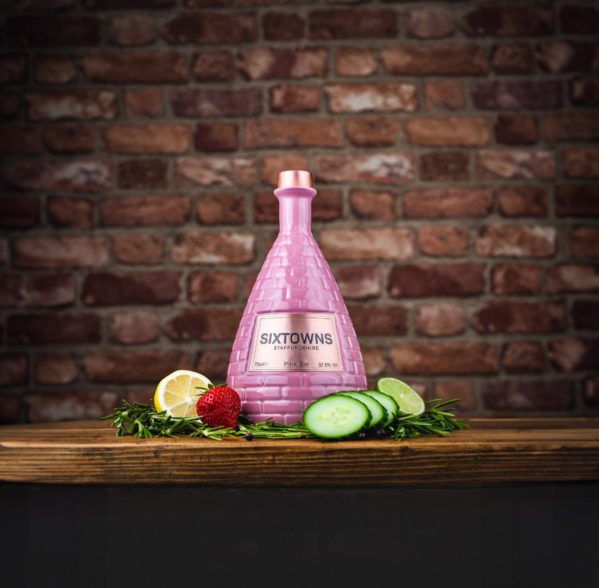 SIXTOWNS Pink Gin bottle with rose gold cap placed on a rustic wooden table, surrounded by cucumber slices, lemon, strawberries, and rosemary, with a brick wall background.