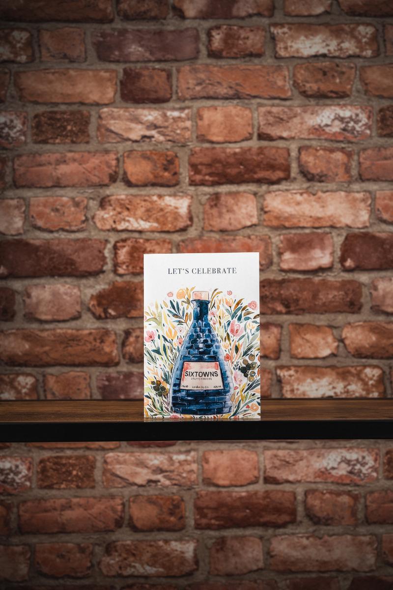 Greeting card featuring a painted SIXTOWNS ceramic bottle with floral artwork and the words "LET’S CELEBRATE", displayed on a rustic wooden shelf in front of a red brick wall.