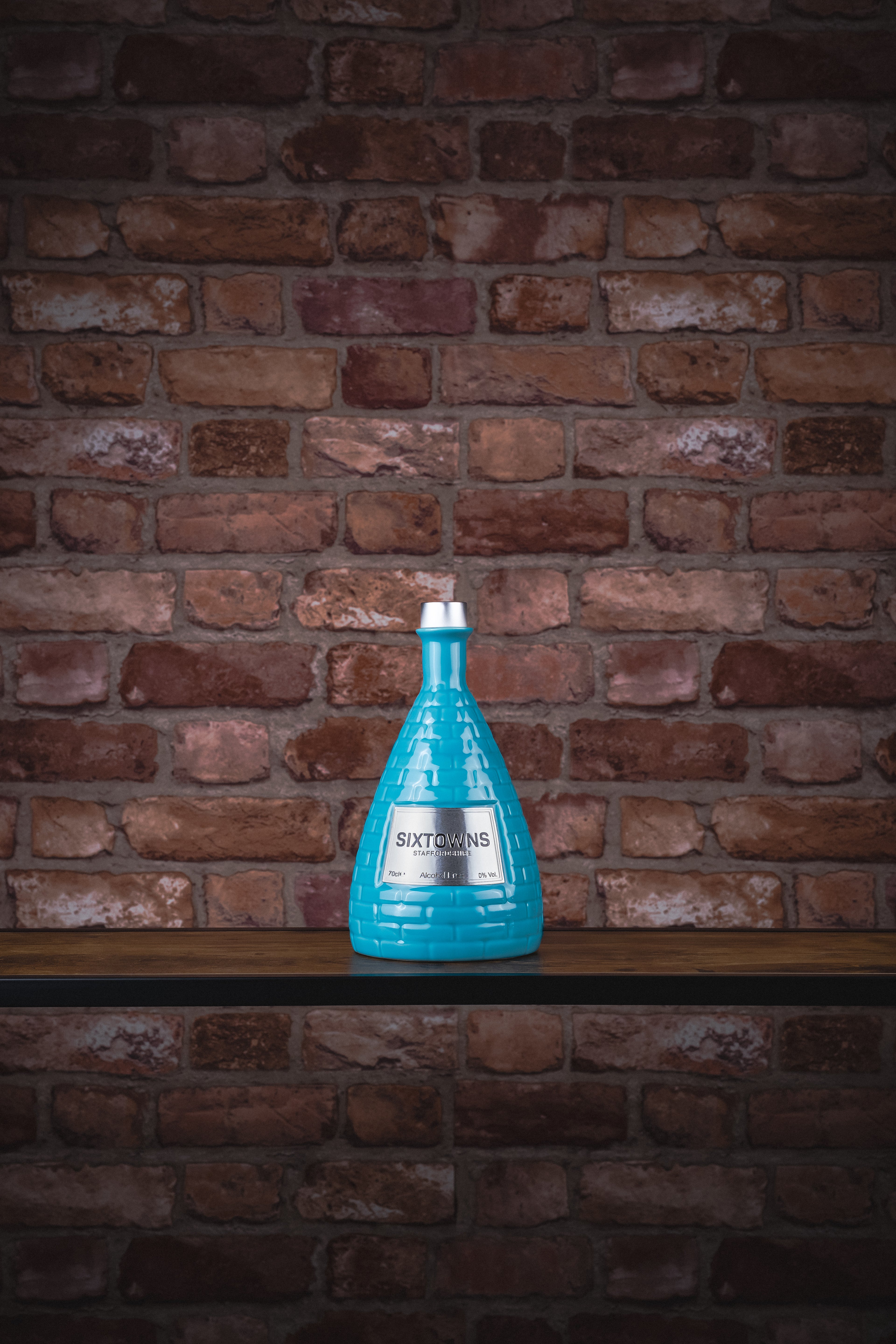 SIXTOWNS Alcohol-Free 0.0% ceramic bottle displayed against a brick wall background, showcasing its signature turquoise kiln-shaped design