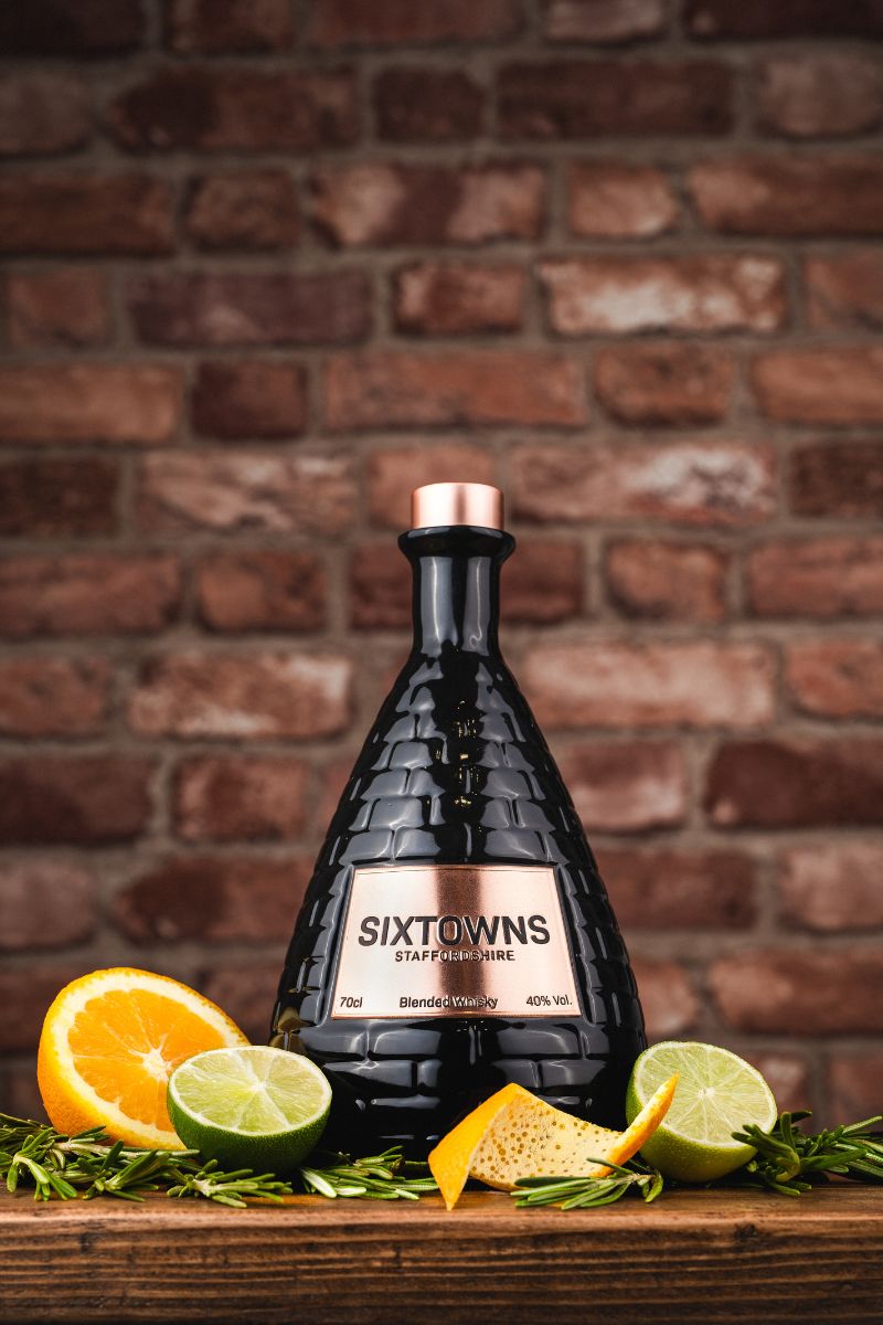 SIXTOWNS Blended Whisky bottle with copper cap displayed on a rustic wooden surface with lime, orange slices, and rosemary, set against a red brick background.