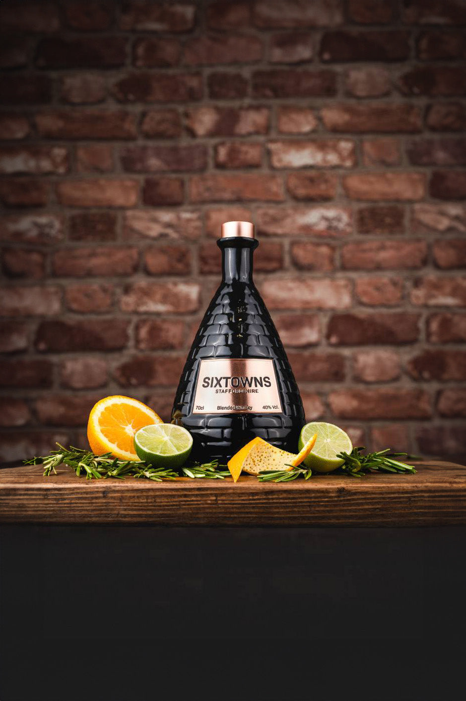 SIXTOWNS Blended Whisky bottle with copper cap displayed on a rustic wooden surface with lime, orange slices, and rosemary, set against a red brick background.