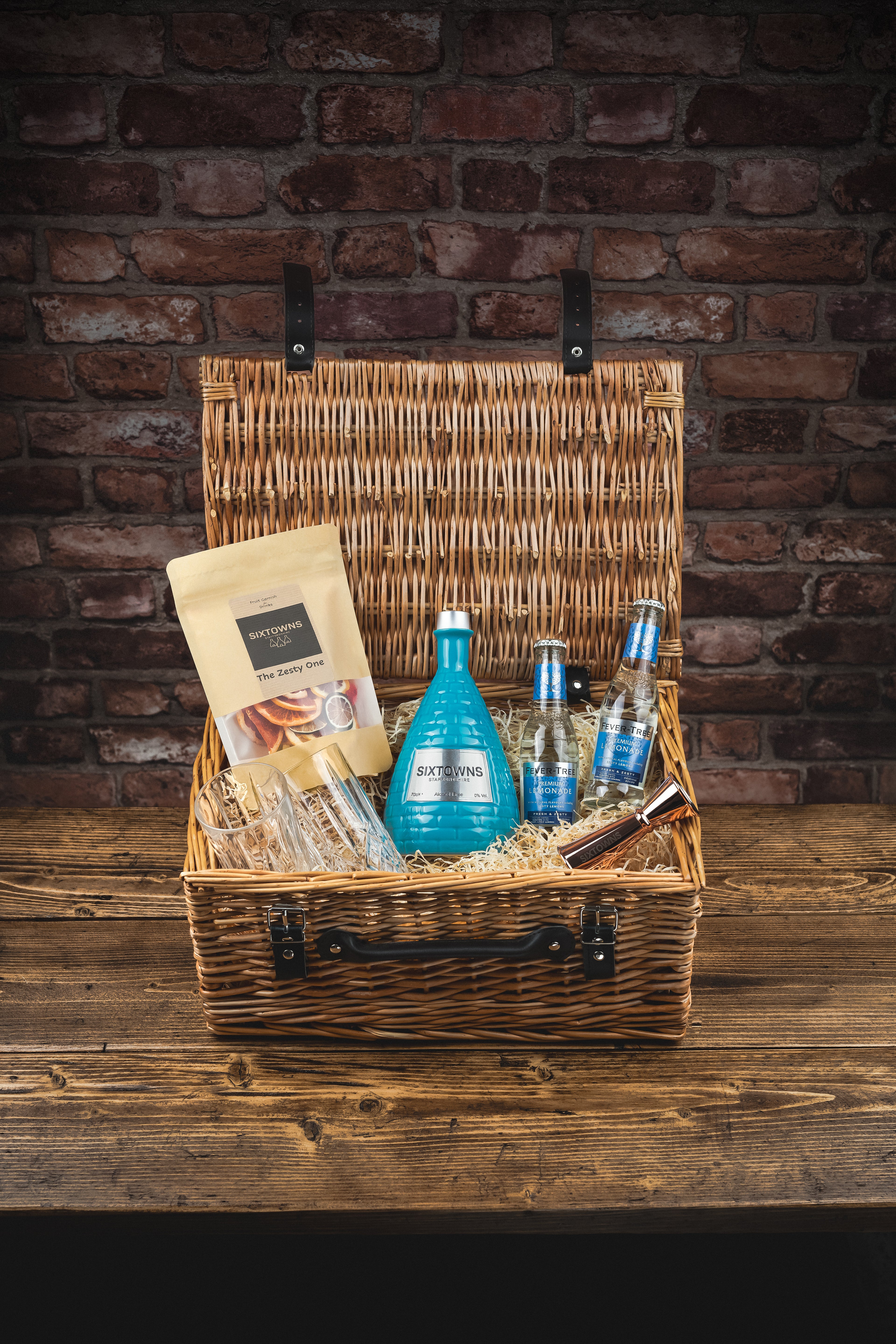 SIXTOWNS Deluxe Hamper gift set featuring alcohol-free spirit, premium mixers, dried fruit garnish, copper jigger and crystal glasses in a wicker basket against a rustic brick wall