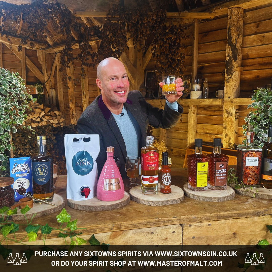 TV presenter smiling and holding a glass of spirit during an ITV showcase featuring a range of British craft spirits, including SIXTOWNS Pink Gin in a ceramic bottle, set against a rustic wooden bar with greenery and artisanal products on display