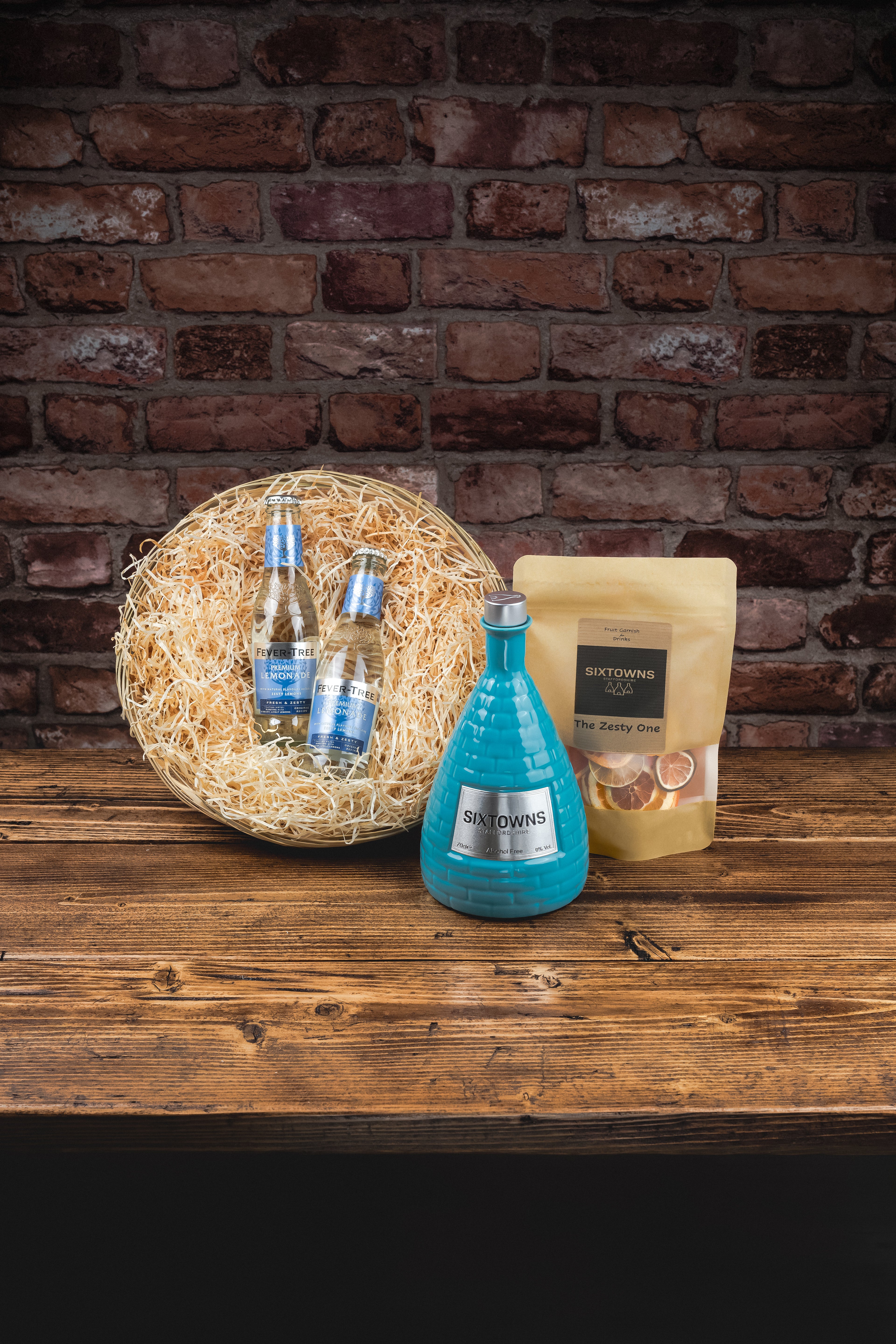 SIXTOWNS Ultimate Hamper gift set featuring alcohol-free spirit, two premium mixers, dried fruit garnish, and round straw-filled hamper tray, styled on rustic wood with brick backdrop